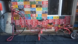 A long, uniquely designed red bicycle with a large front wheel is positioned indoors against a wall covered with various colorful vintage license plates from different regions. It appears to be a multi-seater bike with intricate metalwork and a slightly antiquated look, combining elements of a penny-farthing with modern bicycles.