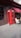 A classic red telephone booth is positioned on a paved sidewalk against the wall of a building. The booth features glass paneling and a sign that reads 'TELEPHONE' at the top. To the right, there is a small outdoor seating area with a metal table and cream-colored chairs, partially shaded by a red awning bearing the word 'WOK'. The brown brick wall and pavement create a neutral backdrop for the vivid red of the telephone booth.