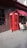 A classic red telephone booth is positioned on a paved sidewalk against the wall of a building. The booth features glass paneling and a sign that reads 'TELEPHONE' at the top. To the right, there is a small outdoor seating area with a metal table and cream-colored chairs, partially shaded by a red awning bearing the word 'WOK'. The brown brick wall and pavement create a neutral backdrop for the vivid red of the telephone booth.