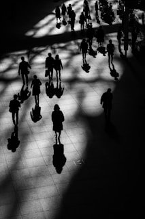 Silhouetted figures walk across a tile floor with dramatic shadows cast by overhead light. The play of light and shadow creates an abstract pattern on the ground, emphasizing the contrast between the illuminated areas and the dark silhouettes. A variety of individuals appear to move in different directions, suggesting a bustling, dynamic environment.