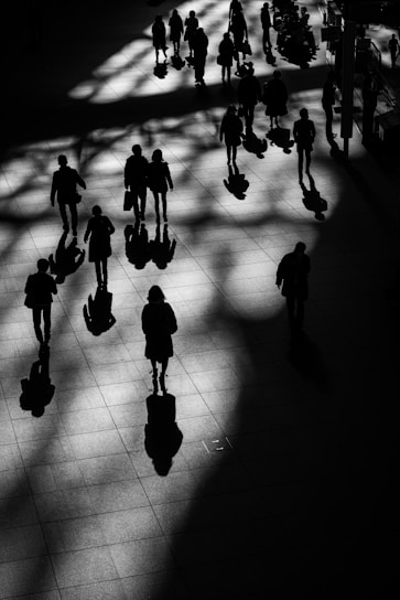 Silhouetted figures walk across a tile floor with dramatic shadows cast by overhead light. The play of light and shadow creates an abstract pattern on the ground, emphasizing the contrast between the illuminated areas and the dark silhouettes. A variety of individuals appear to move in different directions, suggesting a bustling, dynamic environment.