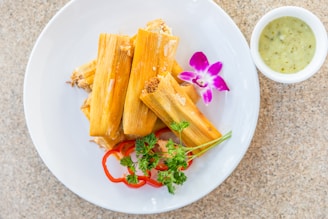 Several tamales are arranged on a white plate, garnished with red bell pepper slices, fresh green parsley, and a purple orchid flower. A small white cup filled with green sauce is placed beside the plate on a light-colored, speckled surface.