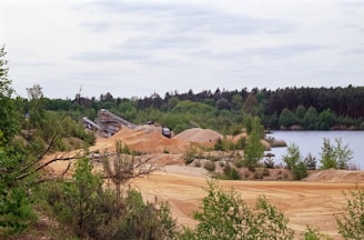 A sand or gravel quarry area with mounds of sand and a conveyor belt system. The scene is surrounded by lush greenery and dense forest. A body of water is visible to the right, creating a contrast with the industrial machinery.