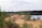 A sand or gravel quarry area with mounds of sand and a conveyor belt system. The scene is surrounded by lush greenery and dense forest. A body of water is visible to the right, creating a contrast with the industrial machinery.