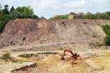 A large excavator is digging earth in a quarry, surrounded by mounds of dirt and sparse vegetation. The landscape includes a steep, eroded hill in the background with some green foliage on top.