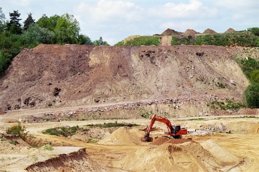 A large excavator is digging earth in a quarry, surrounded by mounds of dirt and sparse vegetation. The landscape includes a steep, eroded hill in the background with some green foliage on top.