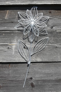 A decorative metal flower structure is mounted on a rustic wooden wall. The flower is intricately designed with thin metal lines forming the petals and leaves, creating a delicate and artistic appearance.