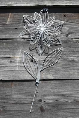 A decorative metal flower structure is mounted on a rustic wooden wall. The flower is intricately designed with thin metal lines forming the petals and leaves, creating a delicate and artistic appearance.