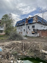 A two-story building with a damaged roof covered in blue tarp. The surrounding area shows an empty, barren yard with scattered debris and a large, shallow pit filled with green water. Sparse vegetation, including some trees with bare branches, can be seen around the building. The sky is overcast with patches of clouds.