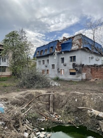 A two-story building with a damaged roof covered in blue tarp. The surrounding area shows an empty, barren yard with scattered debris and a large, shallow pit filled with green water. Sparse vegetation, including some trees with bare branches, can be seen around the building. The sky is overcast with patches of clouds.
