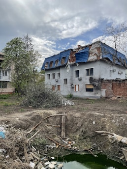 A two-story building with a damaged roof covered in blue tarp. The surrounding area shows an empty, barren yard with scattered debris and a large, shallow pit filled with green water. Sparse vegetation, including some trees with bare branches, can be seen around the building. The sky is overcast with patches of clouds.