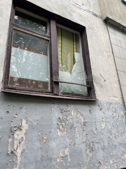 A window with a broken pane is set in a worn-out and peeling wall. The window frame is dark and aged, and curtains can be seen inside. The wall is cracked and the paint is flaking, revealing different layers underneath.