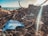 A collection of debris and litter is scattered along the shoreline with sticks, twigs, and various pieces of trash, including a prominent blue plastic fragment. The background features a view of the ocean under a partly cloudy sky with sunlight streaming through.