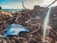 A collection of debris and litter is scattered along the shoreline with sticks, twigs, and various pieces of trash, including a prominent blue plastic fragment. The background features a view of the ocean under a partly cloudy sky with sunlight streaming through.