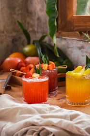 Three glasses filled with colorful fruit drinks, garnished with fresh fruit pieces and leaves, are placed on a wooden table. In the background, there is a tray filled with various fruits including apples and oranges, along with green leaves and a rustic wall with a window.