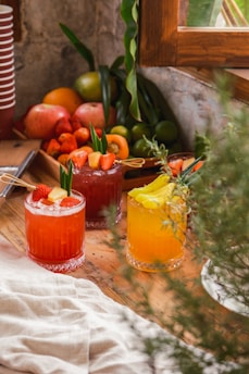 A wooden table is adorned with colorful fruit cocktails garnished with slices of fruit and leaves, placed beside a collection of fresh fruits including strawberries, limes, and apples. A rustic window frame and greenery add to the cozy and inviting atmosphere.