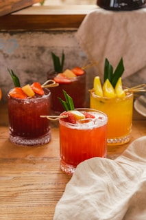 Several glasses filled with colorful cocktails are garnished with fruit and small leaves. The drinks are placed on a wooden table with a light-colored cloth nearby. The background has a rustic appearance, contributing to a cozy and inviting atmosphere.