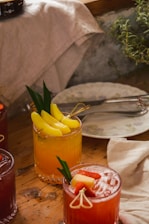 A wooden table is set with various colorful cocktails. A bright yellow drink garnished with pineapple slices and green leaves sits in a faceted glass. Next to it is a red beverage topped with strawberry pieces and a fruit skewer. A light-colored cloth and ceramic plate with floral patterns can be seen in the background along with green foliage, adding a rustic and refreshing atmosphere.