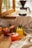 A cozy setting on a wooden table with four vibrant fruit cocktails garnished with slices of pineapple and strawberries. A coffee dripper with a carafe sits near an open window, letting in soft natural light. A beige cloth is draped beside neatly stacked plates and cutlery.
