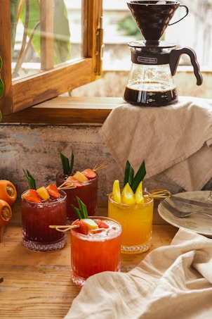 A cozy setting on a wooden table with four vibrant fruit cocktails garnished with slices of pineapple and strawberries. A coffee dripper with a carafe sits near an open window, letting in soft natural light. A beige cloth is draped beside neatly stacked plates and cutlery.