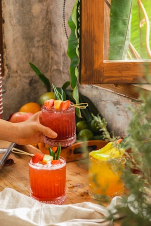A cozy setting with two colorful cocktails on a wooden table, surrounded by fresh fruits like oranges and apples. A hand is reaching out to pick up a red cocktail garnished with strawberries and a pineapple piece, while another glass with a bright yellow liquid garnished with citrus slices is nearby. The background includes lush green leaves and a rustic window frame, creating a fresh and natural ambiance.