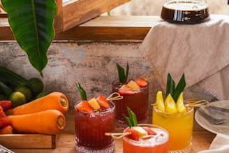 A rustic setting with a variety of fresh juices in glass cups garnished with fruit pieces and leaves. Carrots, limes, and strawberries are neatly arranged next to the drinks. A coffee carafe sits on a wooden counter beside a white cloth.