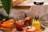 A rustic setting with a variety of fresh juices in glass cups garnished with fruit pieces and leaves. Carrots, limes, and strawberries are neatly arranged next to the drinks. A coffee carafe sits on a wooden counter beside a white cloth.