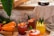 A rustic setting with a variety of fresh juices in glass cups garnished with fruit pieces and leaves. Carrots, limes, and strawberries are neatly arranged next to the drinks. A coffee carafe sits on a wooden counter beside a white cloth.