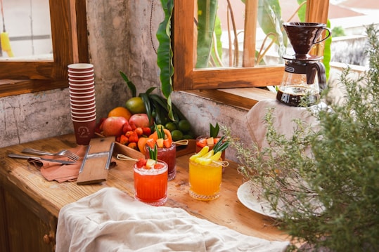 A cozy kitchen scene with two colorful drinks garnished with fruit and herbs placed on a wooden counter. A stack of paper cups, various fruits including apples, strawberries, and citrus, and a coffee maker are nearby. Greenery and wood elements add a natural touch.