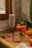 A rustic kitchen counter with a stack of red paper cups next to a wooden window frame. Fresh fruits like strawberries, apples, mangoes, and oranges are arranged beside the cups. Silver forks rest on a folded brown cloth napkin, and a wooden mandoline slicer is placed nearby. Two glass jars with red smoothies, garnished with whole strawberries, complete the cozy kitchen setting.