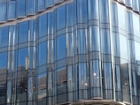 A modern building facade made primarily of large, reflective glass panels with a wavy, undulating design. The glass panels reflect the surrounding environment, including the sky and nearby structures.