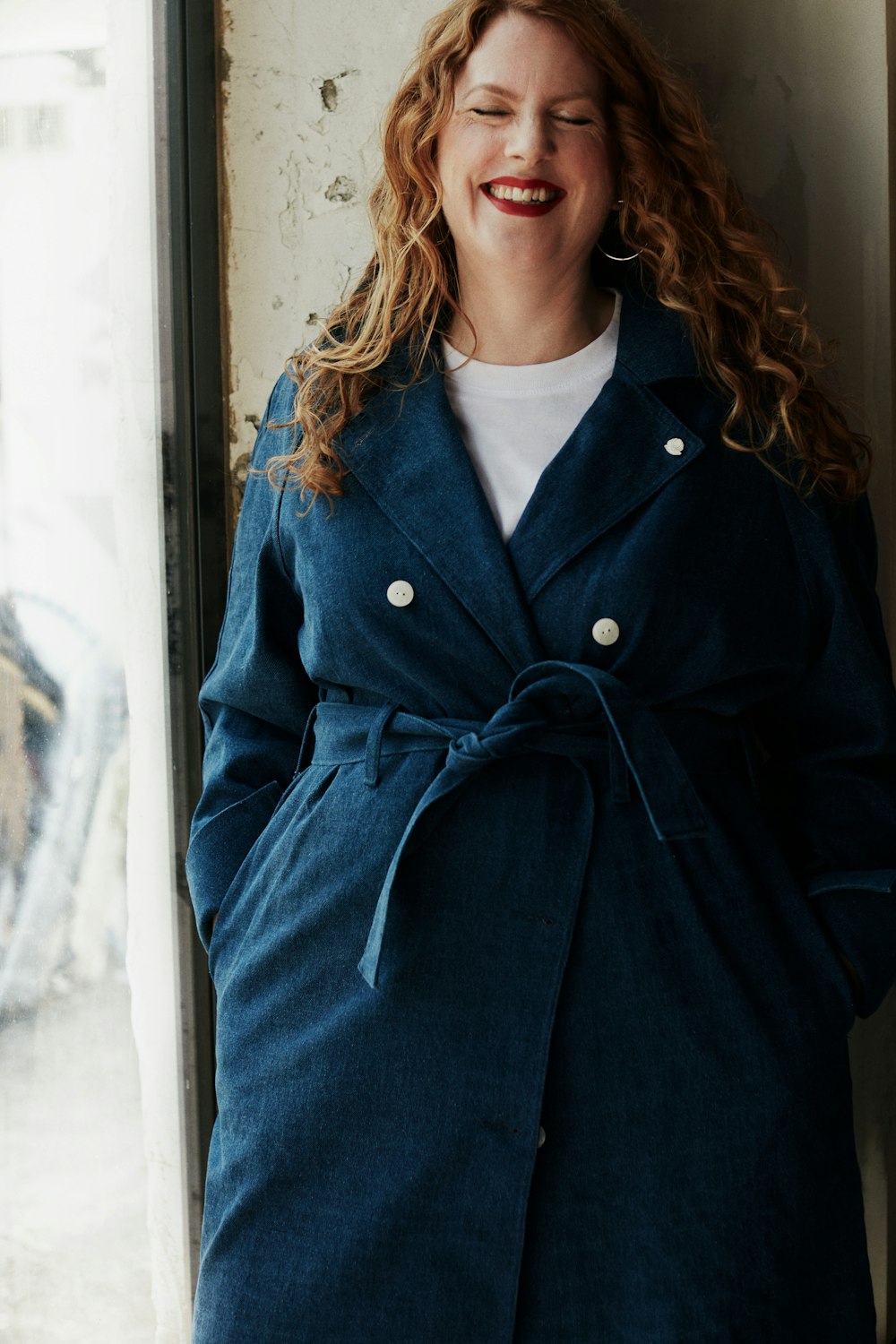 a woman standing next to a window wearing a blue coat