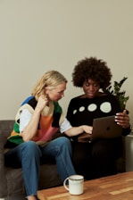 two women sitting on a couch looking at a laptop