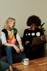 Two people are sitting on a sofa. One is holding a laptop and they both appear to be smiling at the screen. A coffee mug is placed on a wooden table in front of them. The atmosphere seems casual and friendly.