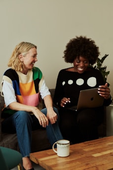Two people are sitting on a sofa. One is holding a laptop and they both appear to be smiling at the screen. A coffee mug is placed on a wooden table in front of them. The atmosphere seems casual and friendly.