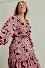 A person wearing a long-sleeve dress with a pink and black floral pattern. The dress has a wrap style, and the person appears confident with a slight smile. The background is plain and neutral, enhancing the focus on the clothing.