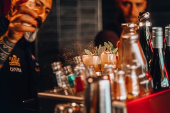 A bartender with tattooed arms is preparing cocktails, adding a final touch or spray to a glass with mint garnish. Various bottles and cocktail shakers are visible in the foreground, indicating a busy bar scene with skillful mixing.
