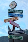 Several colorful directional signs point to various locations such as a prayer room, toilet, parking, Pincuran Tujuh, souvenir and culinary kiosk, and exit. The signs are set against a blue sky and a mountainous backdrop, featuring a traditional Minangkabau house roof in the distance. An emblem at the top reads 'Tanah Datar Authentic Minangkabau.'