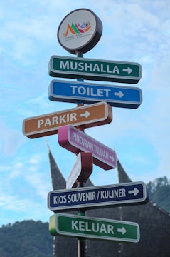 Several colorful directional signs point to various locations such as a prayer room, toilet, parking, Pincuran Tujuh, souvenir and culinary kiosk, and exit. The signs are set against a blue sky and a mountainous backdrop, featuring a traditional Minangkabau house roof in the distance. An emblem at the top reads 'Tanah Datar Authentic Minangkabau.'