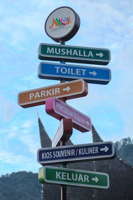 Several colorful directional signs point to various locations such as a prayer room, toilet, parking, Pincuran Tujuh, souvenir and culinary kiosk, and exit. The signs are set against a blue sky and a mountainous backdrop, featuring a traditional Minangkabau house roof in the distance. An emblem at the top reads 'Tanah Datar Authentic Minangkabau.'