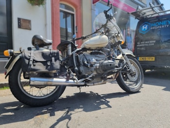 A vintage motorcycle with a sidecar is parked on a street next to a black van. The motorcycle has a large saddlebag and a windshield. The van has signage with contact information and a logo for a property services company. The scene is set in front of a building with a red and white facade, and the lighting suggests it is a sunny day.