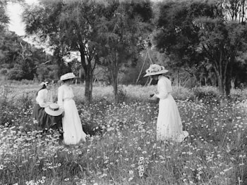 Three women in vintage clothing explore a field of wildflowers, surrounded by tall trees. One woman stands holding an object, while the other two kneel among the flowers. The scenery is serene, set in a lush, peaceful natural environment.
