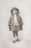 A young child with curly hair stands wearing a vintage outfit. The child is dressed in a coat with large buttons and a fringe scarf, paired with gloves, ankle socks, and shiny shoes. A wide-brimmed hat rests on the child's head, and they appear relaxed, with a neutral expression against a plain background.