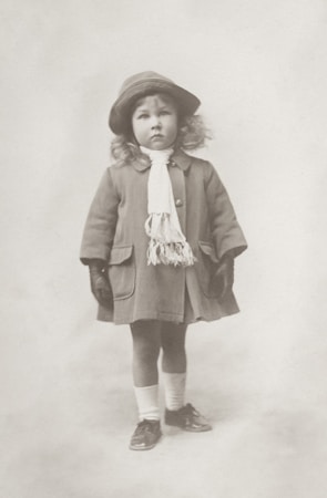 A young child with curly hair stands wearing a vintage outfit. The child is dressed in a coat with large buttons and a fringe scarf, paired with gloves, ankle socks, and shiny shoes. A wide-brimmed hat rests on the child's head, and they appear relaxed, with a neutral expression against a plain background.
