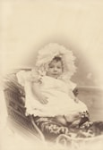 A baby is dressed in a delicate lace bonnet and a flowing white gown, sitting in an ornately designed wicker baby carriage with intricate patterns on the side. The image is in sepia tones, giving a vintage and nostalgic aesthetic.
