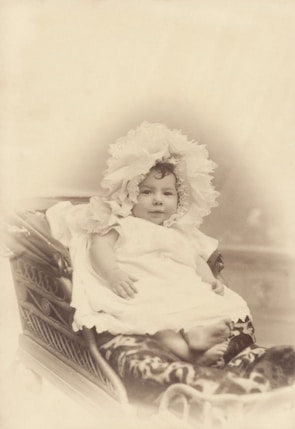 A baby is dressed in a delicate lace bonnet and a flowing white gown, sitting in an ornately designed wicker baby carriage with intricate patterns on the side. The image is in sepia tones, giving a vintage and nostalgic aesthetic.
