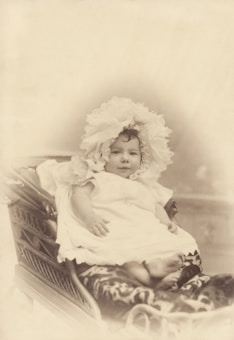 A baby is dressed in a delicate lace bonnet and a flowing white gown, sitting in an ornately designed wicker baby carriage with intricate patterns on the side. The image is in sepia tones, giving a vintage and nostalgic aesthetic.