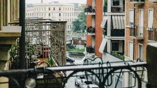 A cityscape view from a balcony overlooking a street with several cars. The foreground includes an ornate metal railing with potted plants. In the background, multi-story buildings with balconies and awnings are visible, along with a large, round, beige structure in the distance.