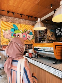 A person wearing a headscarf and striped shirt stands at a cafe counter with a colorful mural in the background. Modern pendant lights hang from the wooden ceiling, and the counter features a large, orange coffee machine. The brick walls and patterned tiles add a cozy, artistic ambiance.