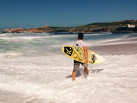 Surfer carrying a board along a rugged Baja beach with crashing waves.